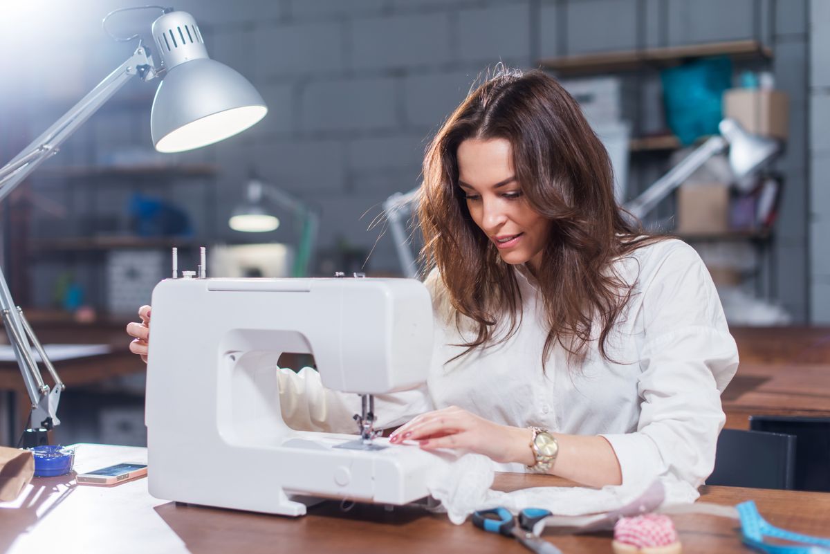 Attractive Caucasian seamstress working stitching with sewing machine at her workplace in studio loft interior