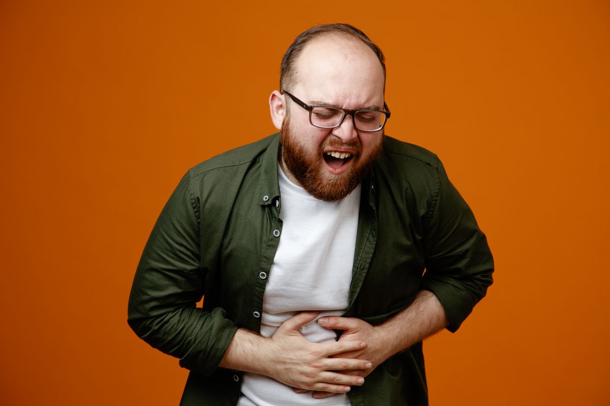 bearded man in casual clothes wearing glasses looking unwell holding hands on his belly feeling pain standing over orange background