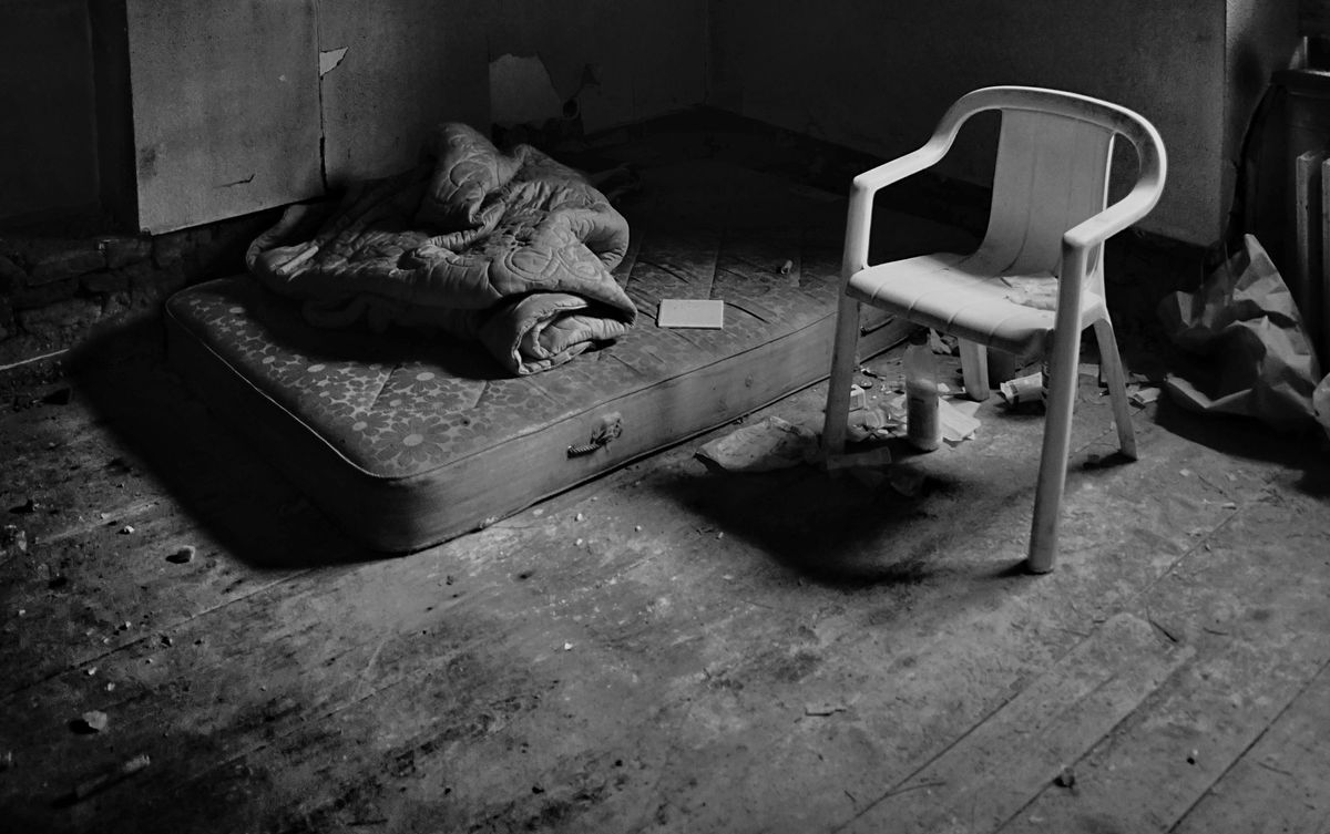 Vertical grayscale shot of the inside of a poor room with a single chair and a mattress