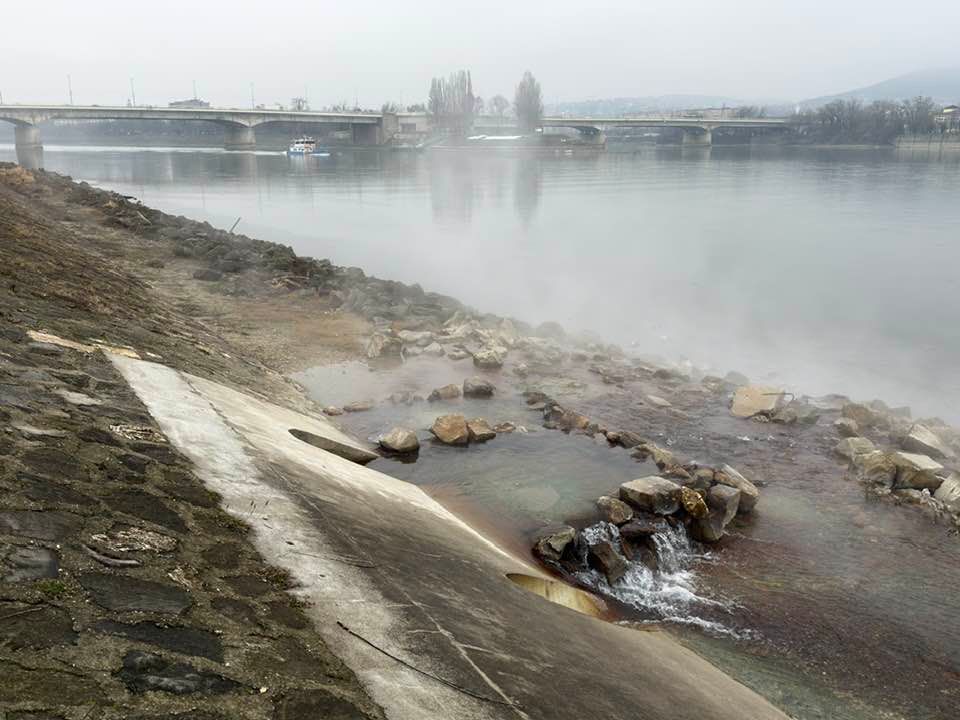 Fent a Duna, távoli, kopasz fák. Elől sziklákkal négyzetalakban kirakott alacsony falsor, amibe gözőlgő víz folyik. 