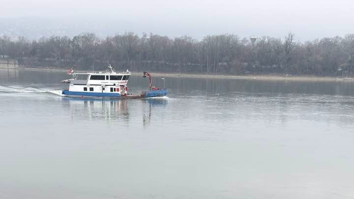 Kék-fehér színű hajó halad a téli Duna angyalföldi szakaszán. Háttérben lomhullott, kopasz fák. 