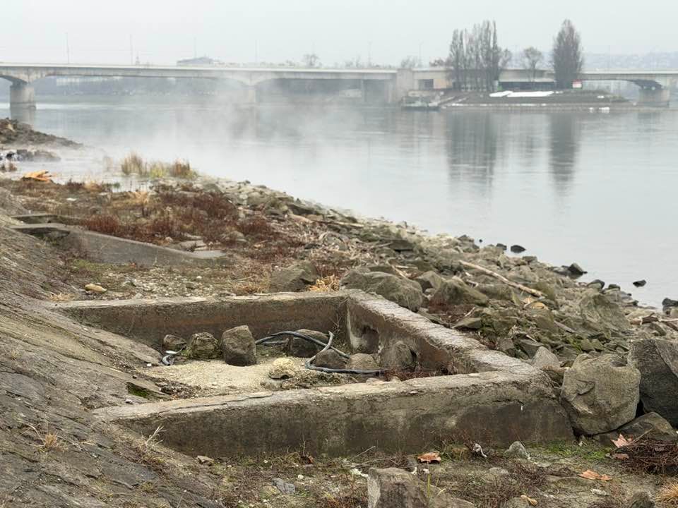 Négyzet alakú, körbe betonozott mélyedés, amiben egy fekete színű gumicső van. Körülötte sziklák, mellette a Duna és a távolban az Árpád híd. 