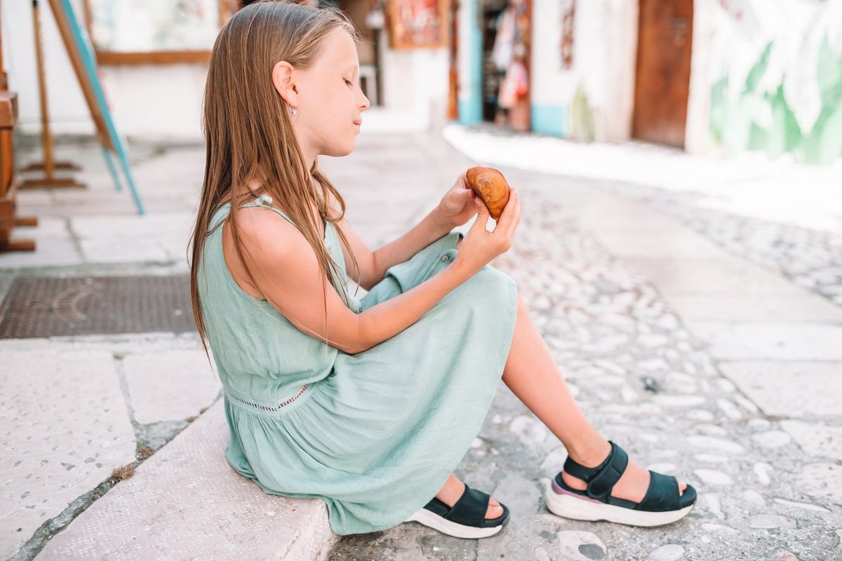 Little girl eats croissant on old streets in european village